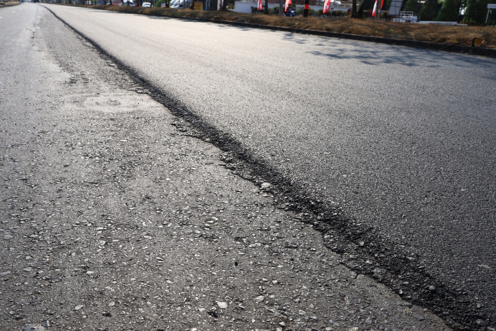 An asphalt road showing a distinct dividing line where a fresh, smooth layer of dark asphalt meets an older, rougher, and worn-down road surface.
