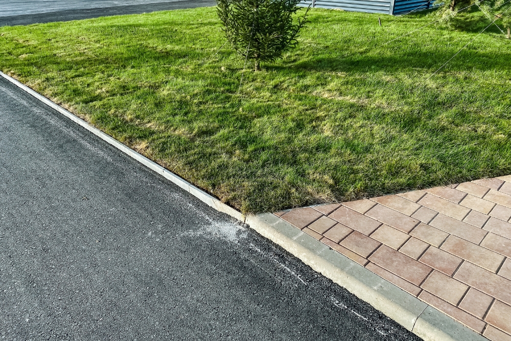 A newly paved asphalt surface bordering a stone curb, a green lawn, and a section of interlocking brick pavers.