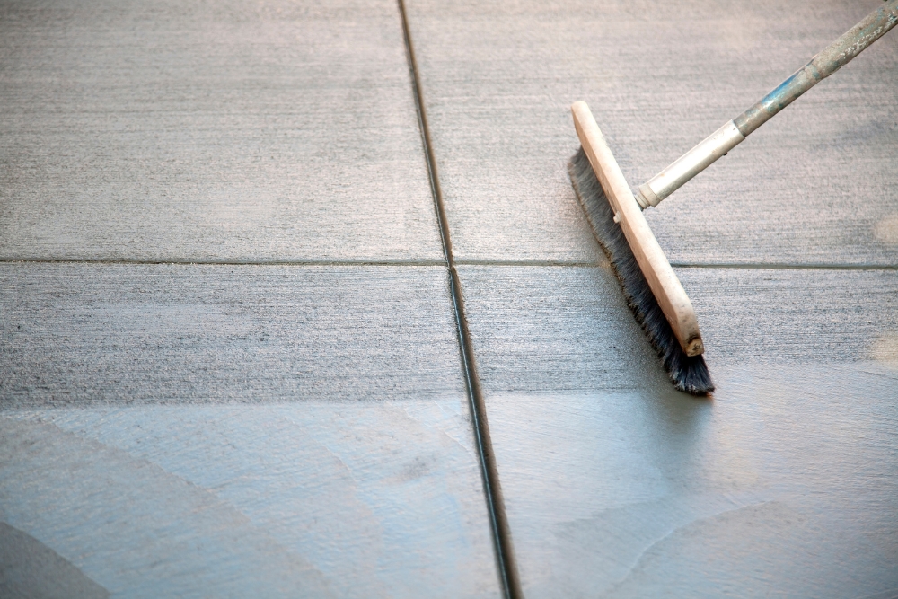 A close-up view of a broom being used to create a textured finish on freshly laid or wet grey concrete slab panels.