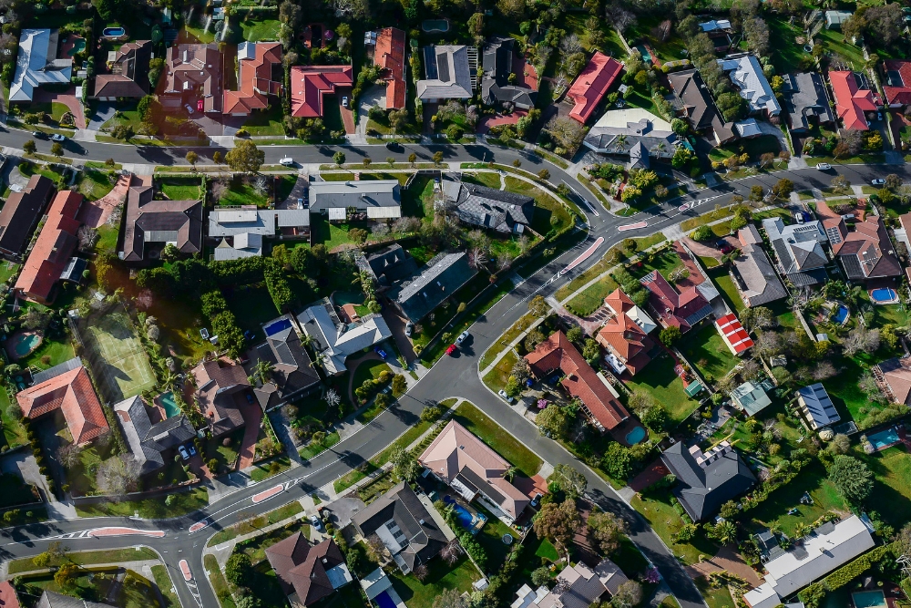 An aerial bird's-eye view of a suburban neighborhood showing houses with red, brown, and grey roofs, green lawns, backyard swimming pools, and winding streets.